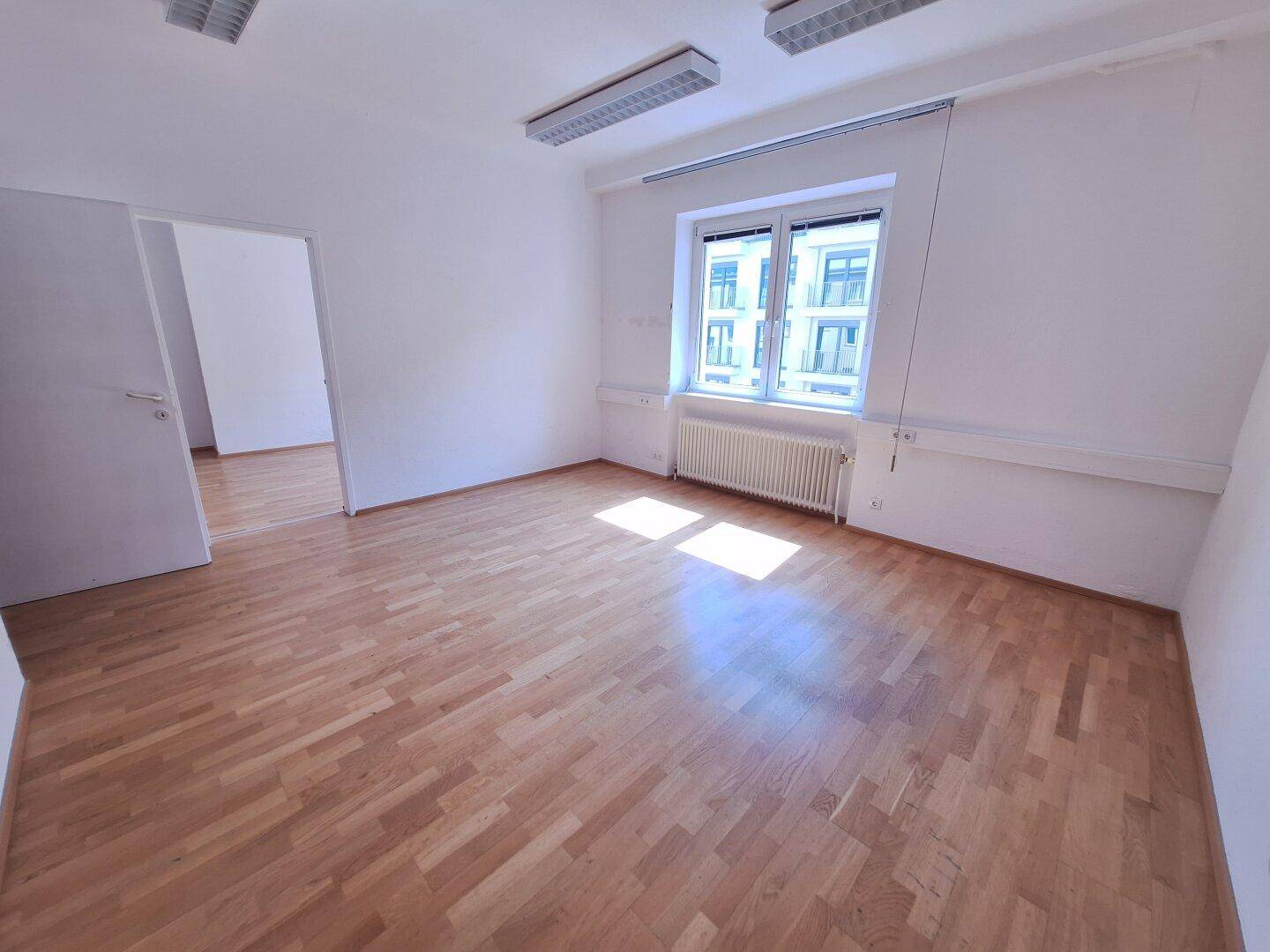Heller Büroraum mit Parkettboden und einem Fenster, das viel Tageslicht hereinlässt und eine angenehme Arbeitsatmosphäre schafft.