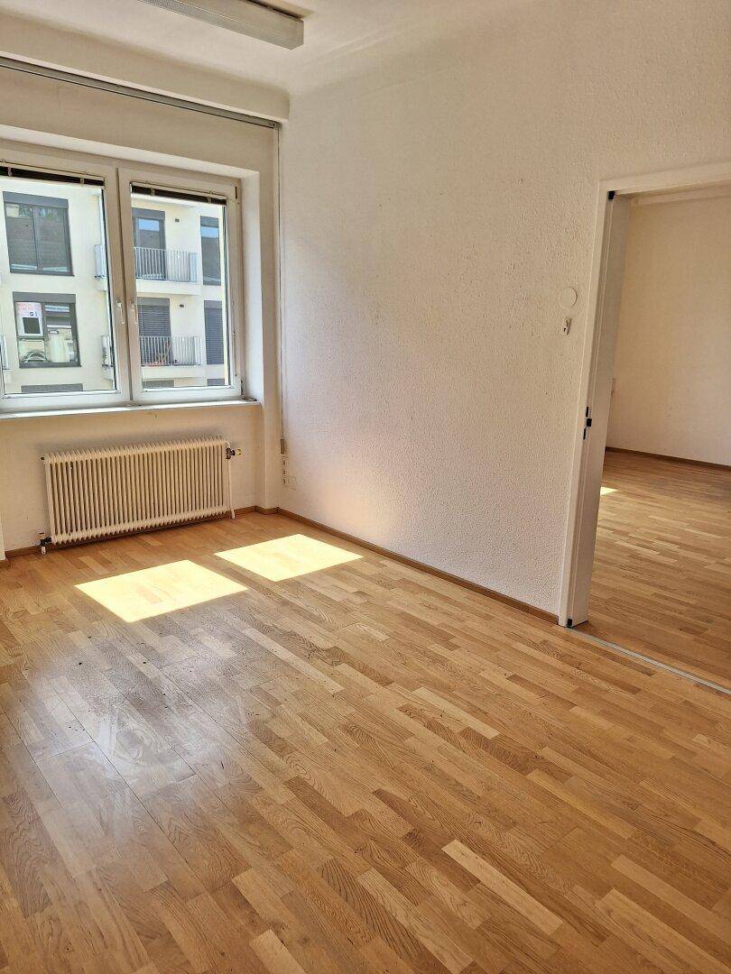 Büroraum mit hellem Parkettboden und einem großen Fenster, das einen Blick auf die umliegenden Gebäude bietet.