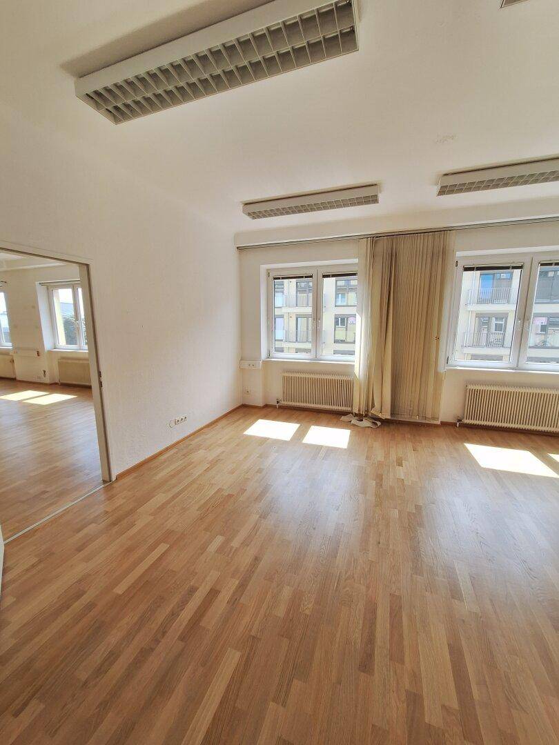 Heller Büroraum mit Holzparkett, großen Fenstern und Vorhängen, die eine angenehme Atmosphäre schaffen.