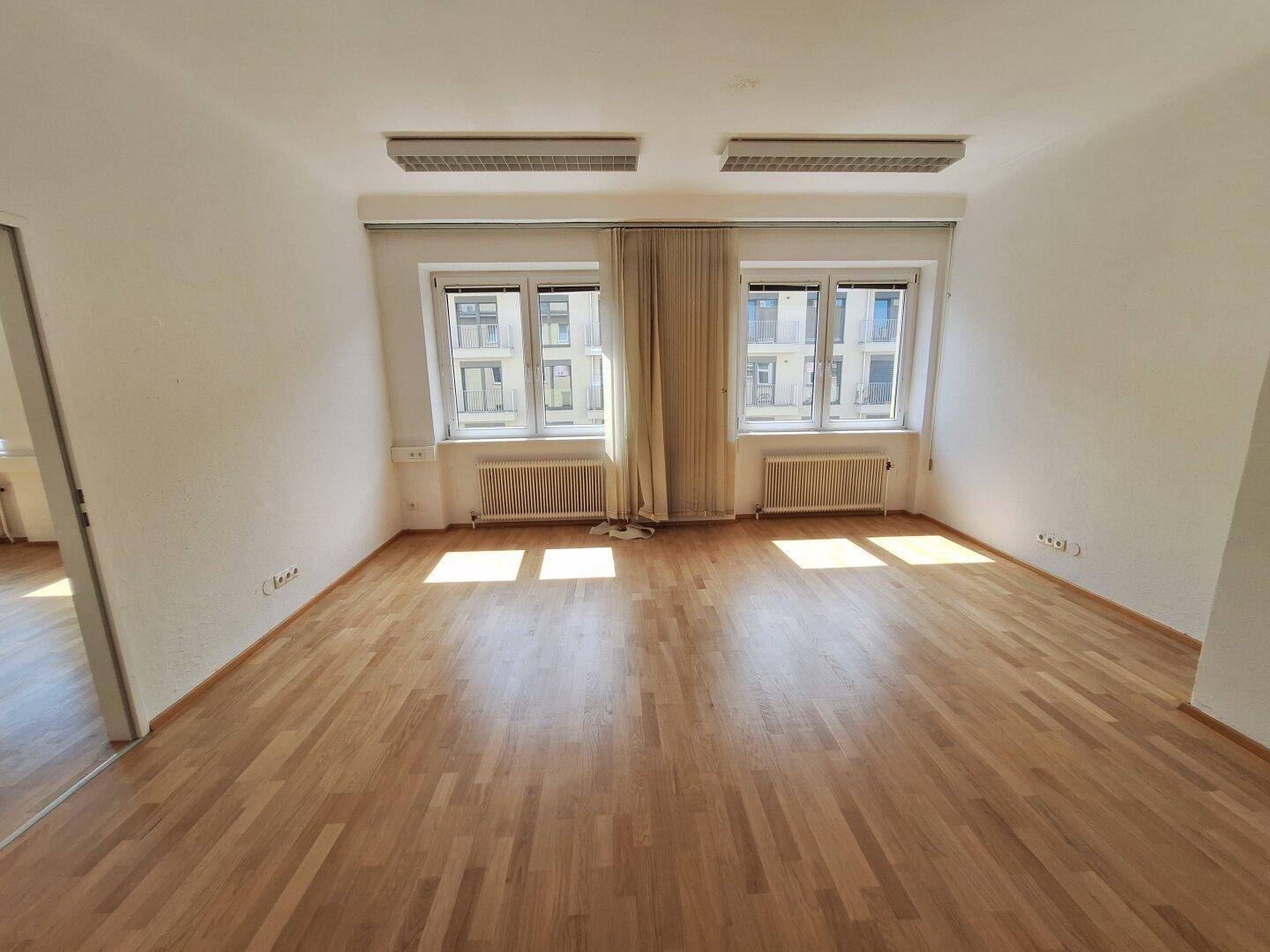 Leerer Büroraum mit zwei großen Fenstern, die viel Sonnenlicht hereinlassen und einen Blick nach draußen bieten.
