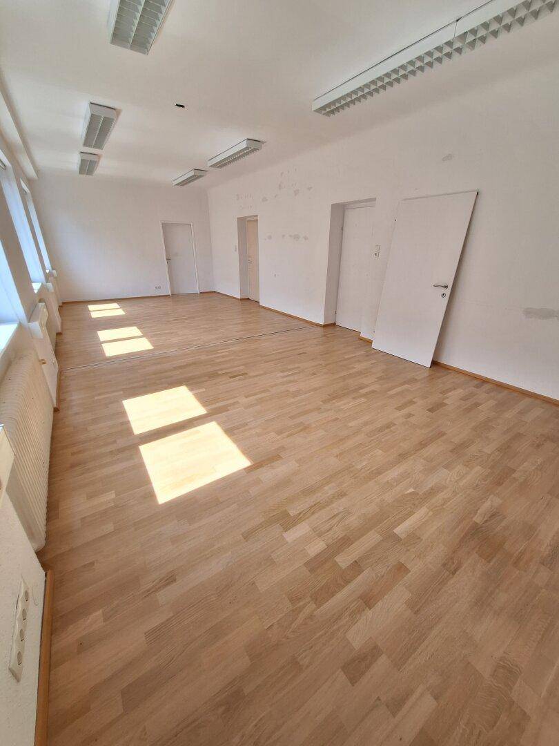Geräumiger Büroraum mit hellem Parkettboden und mehreren Fenstern, die viel Tageslicht hereinlassen.