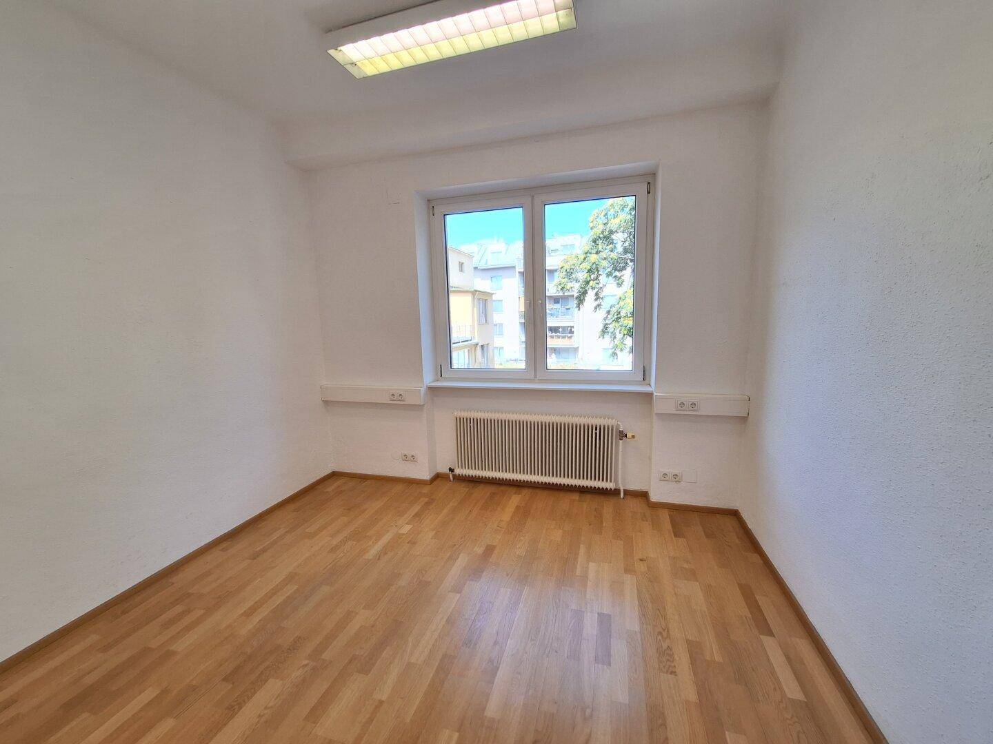 Geräumiges Büro mit Holzparkettboden, Deckenleuchte und einem Fenster mit Blick auf die umliegenden Gebäude.