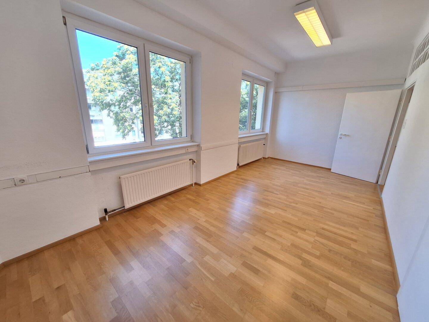 Heller Büroraum mit Blick ins Grüne durch große Fenster, ausgestattet mit Holzparkett und Deckenbeleuchtung.