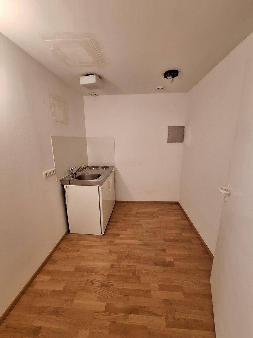 Kleine Küchenzeile in einem schlichten Raum mit Spüle, Kochfeld und Kühlschrank, ideal für eine Büroküche.