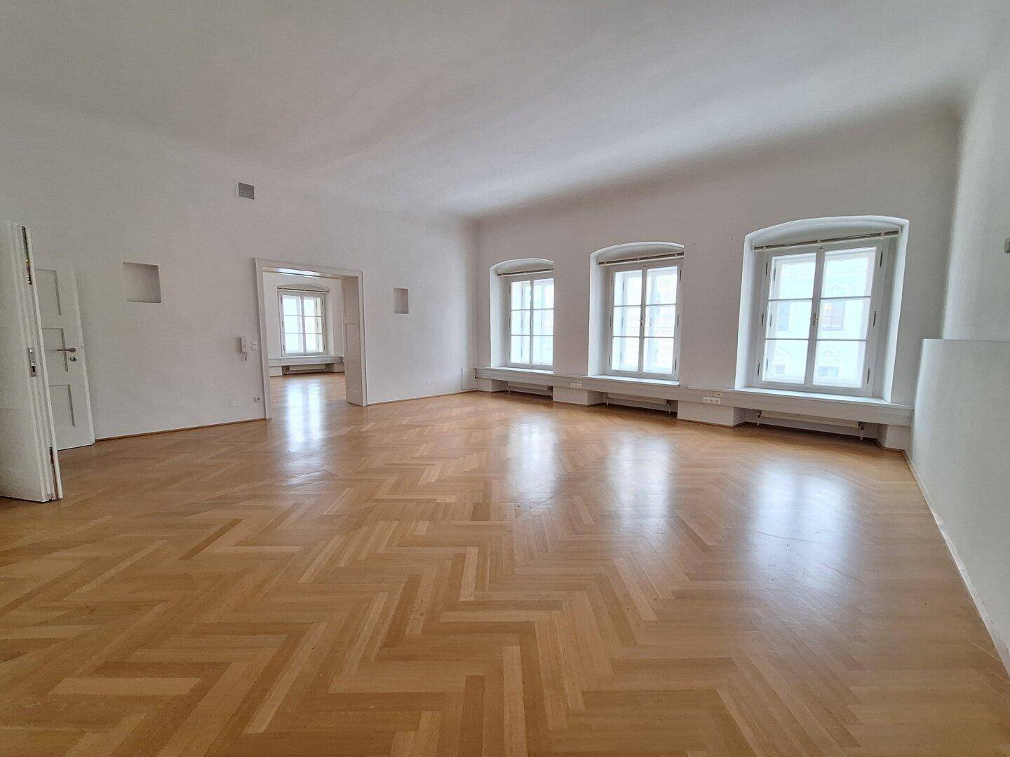 Weitläufiger Raum mit Fischgrätparkett und hohen Fenstern, der durch offene Türen weitere Zimmer verbindet.