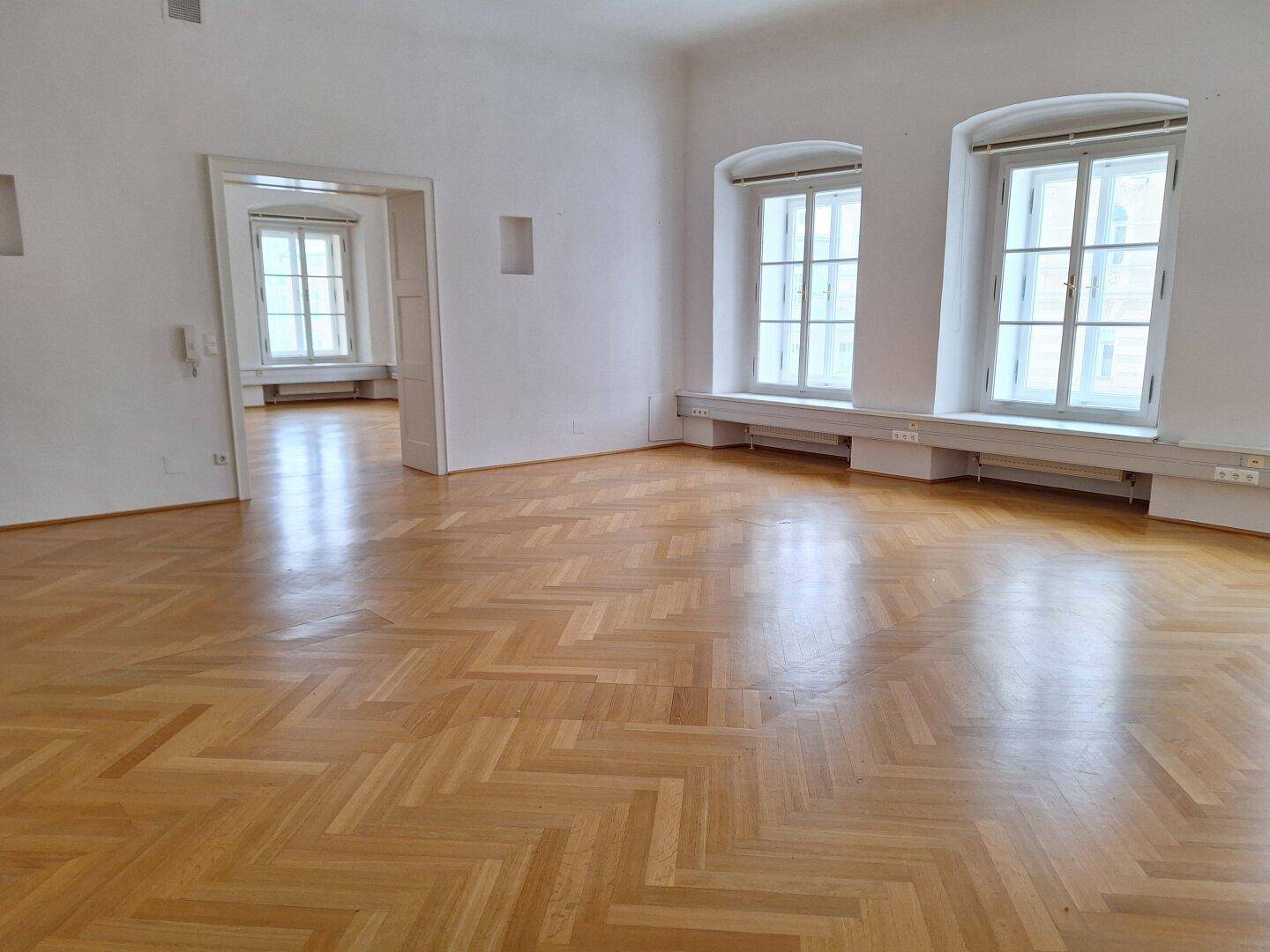 Heller Raum mit glänzendem Fischgrätparkett und hohen Fenstern, die den Blick nach draußen freigeben.
