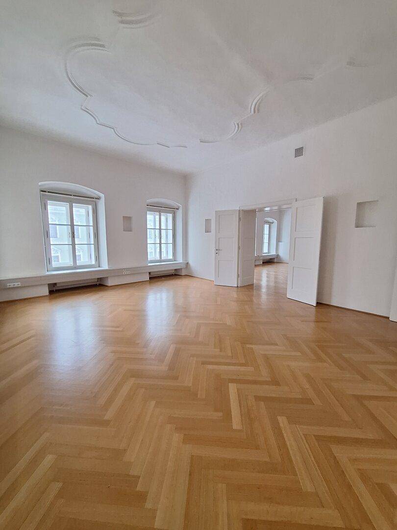 Geräumiges Zimmer mit Fischgrätparkett, hohen Decken und Stuckverzierungen, durch große Fenster hell erleuchtet.