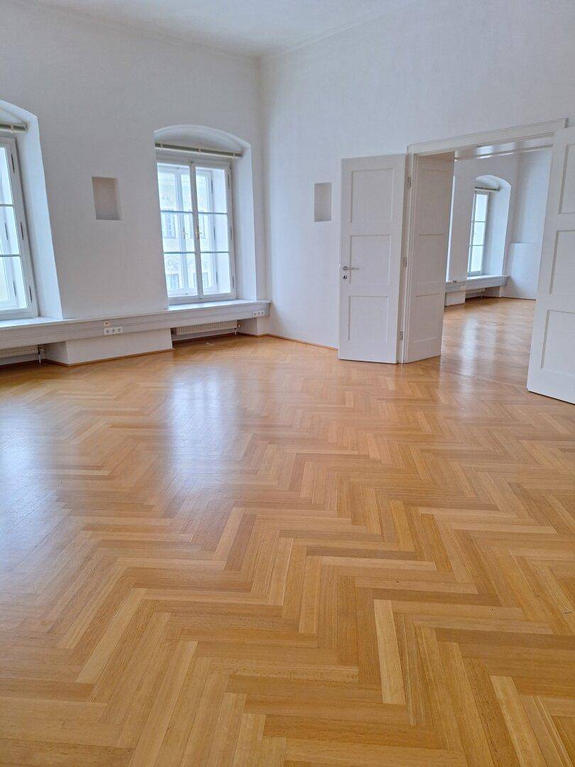 Blick über den glänzenden Fischgrätparkettboden zu den hohen Fenstern, die viel Tageslicht hereinlassen.