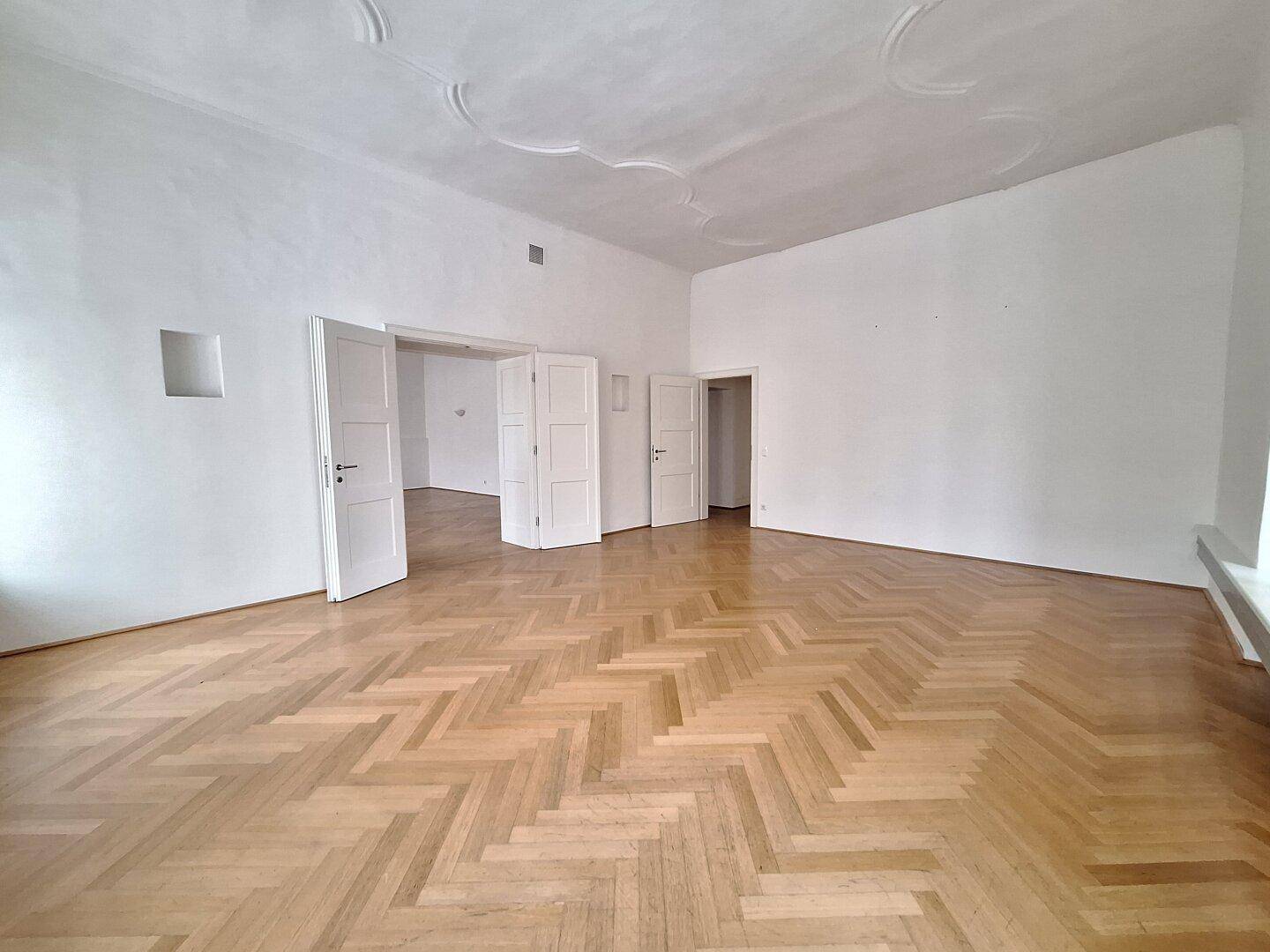 Weitläufiges Zimmer mit Fischgrätparkett und hohen Decken, durch mehrere Türen mit angrenzenden Räumen verbunden.