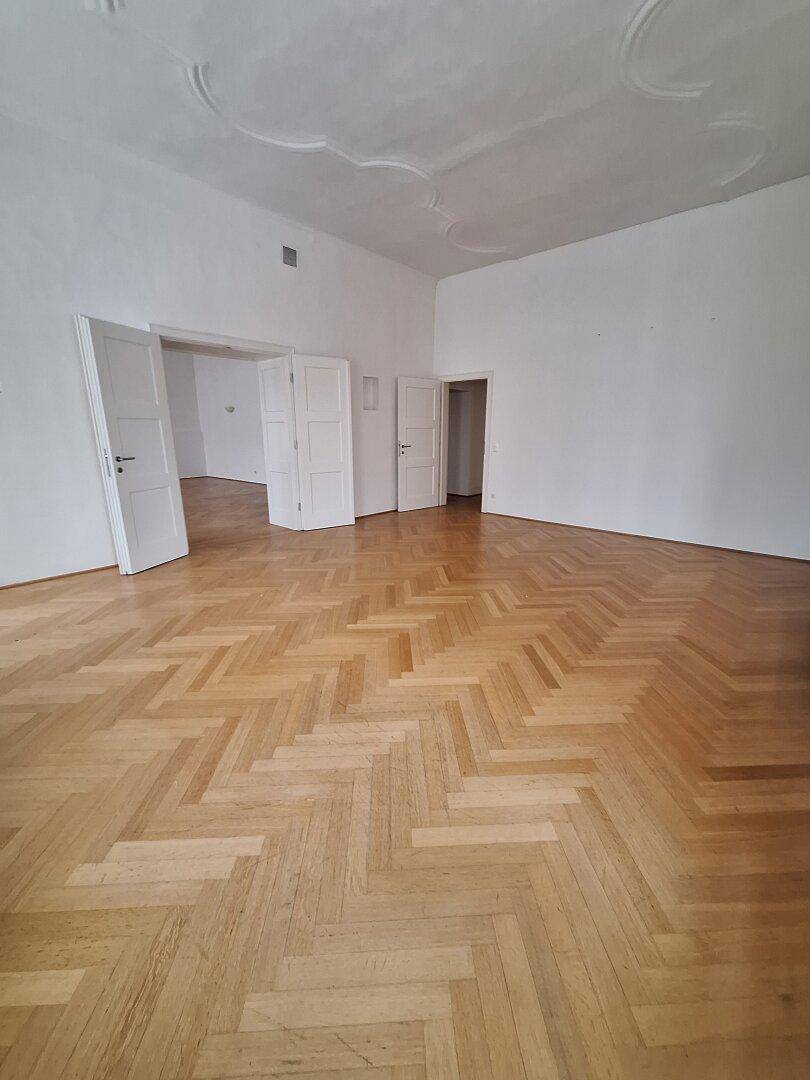 Großes Zimmer mit Fischgrätparkett und hohen Decken, das durch Doppeltüren mit anderen Bereichen verbunden ist.