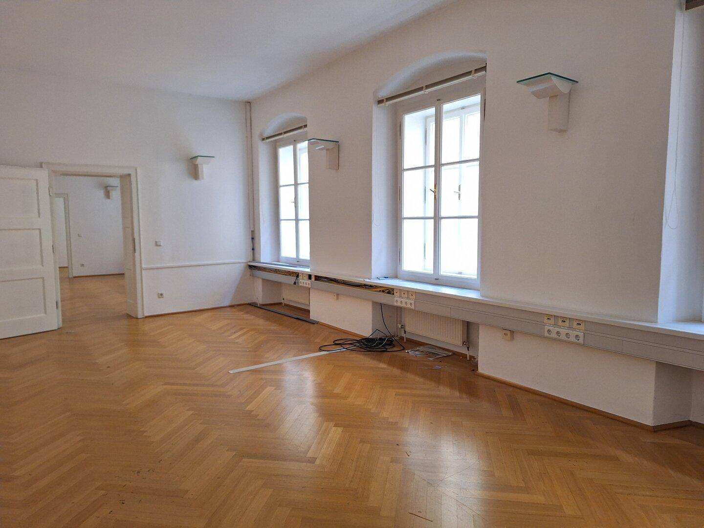 Detailansicht der Fensterfront mit Heizkörpern und Fischgrätparkett in einem hellen, geräumigen Zimmer.
