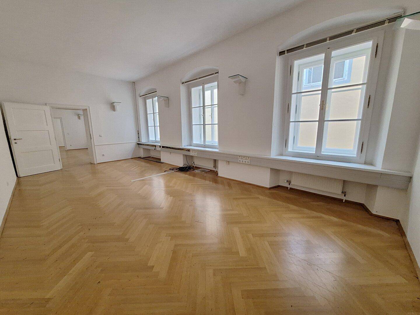 Heller Wohnraum mit drei hohen Fenstern, Fischgrätparkett und Zugang zu weiteren Zimmern.