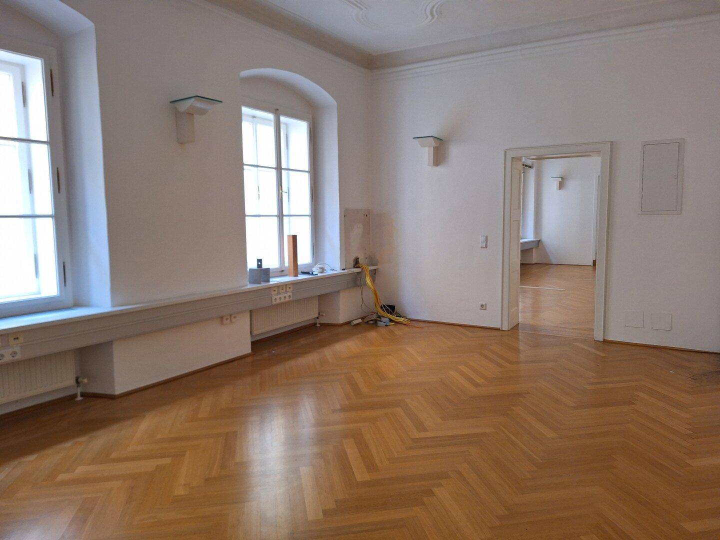 Detailansicht der Fensterfront mit Heizkörpern, Steckdosen und Fischgrätparkett in einem hellen Raum.