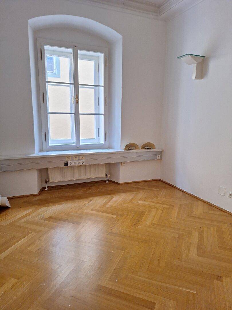 Helle Zimmerecke mit einem hohen Fenster, Fischgrätparkett und einer Wandleuchte, die den Raum erhellt.