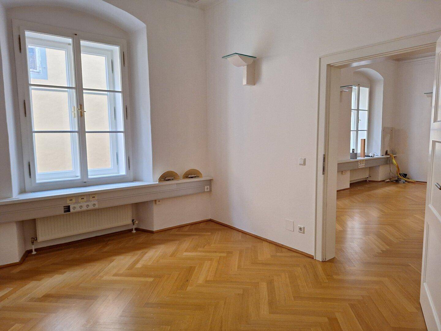 Zimmerecke mit Fischgrätparkett, einem hohen Fenster und einem offenen Durchgang zu einem weiteren Raum.