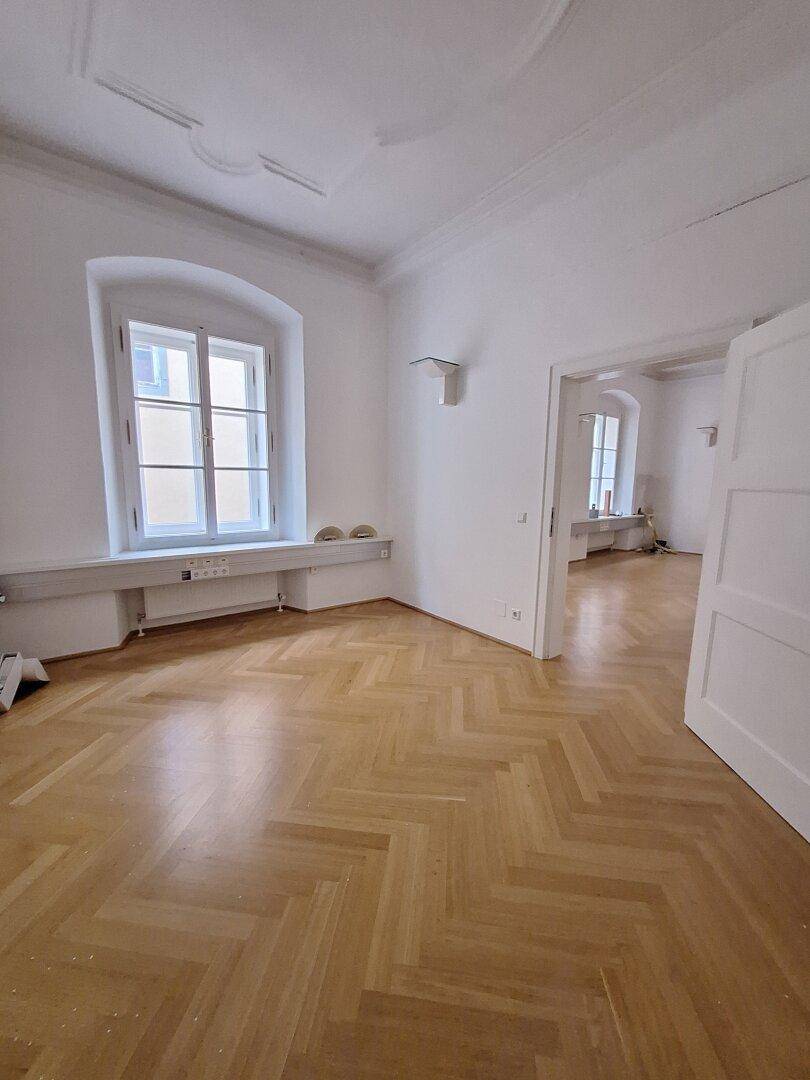 Helle Zimmerecke mit Fischgrätparkett, einem hohen Fenster und einem offenen Durchgang zu einem weiteren Raum.