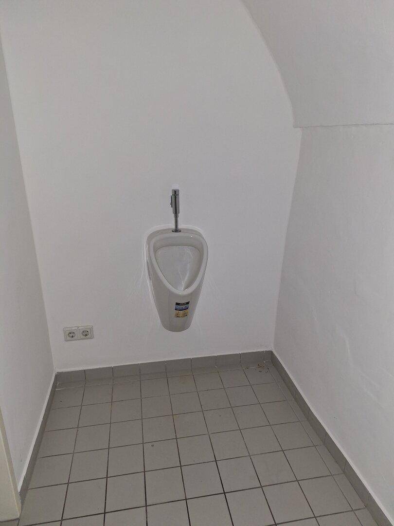 Kleiner Toilettenraum mit Urinal und gefliestem Boden unter einer gewölbten Decke.