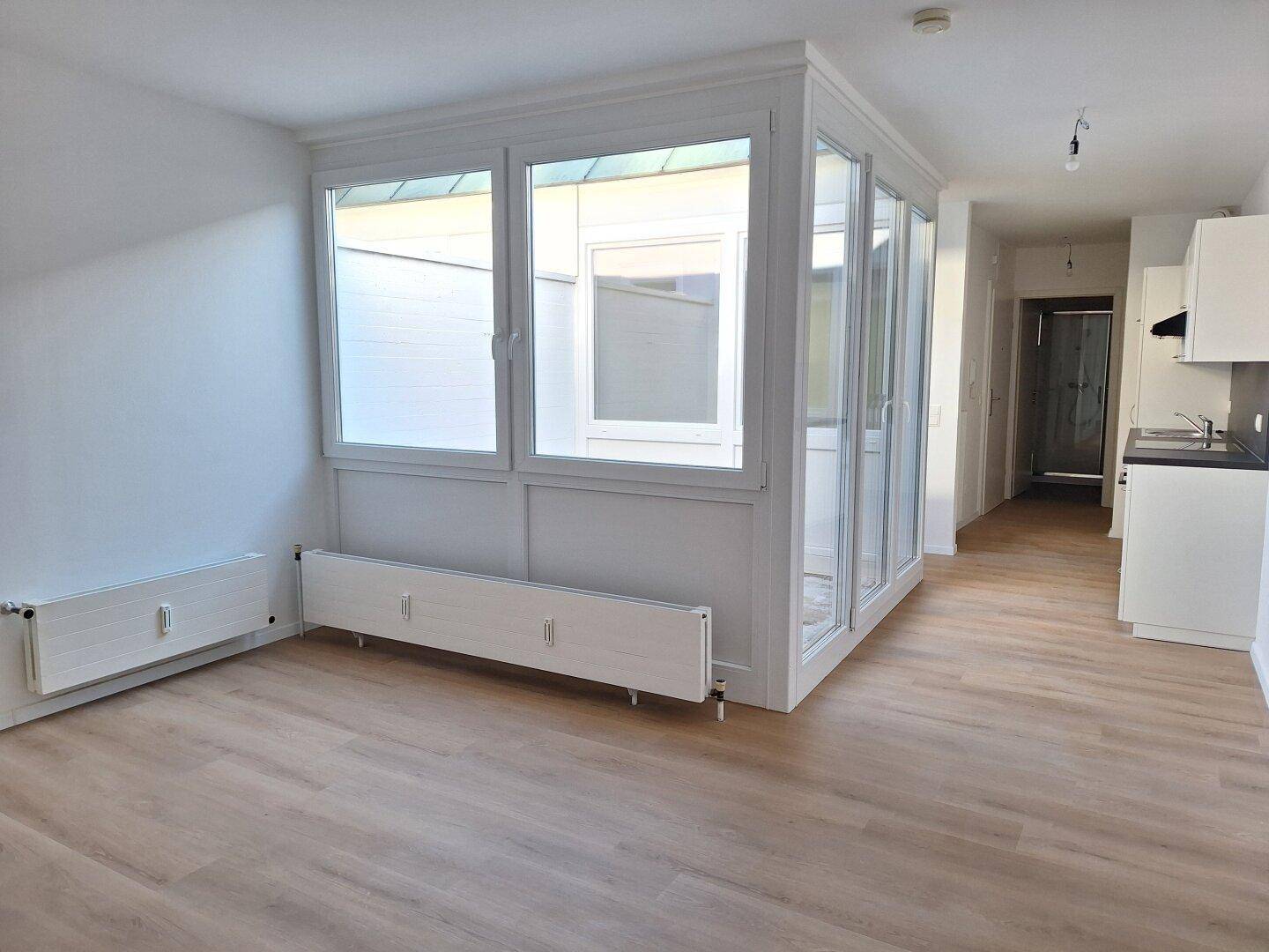 Offener Wohnbereich mit Laminatboden, weißen Wänden und Zugang zu einem verglasten Atrium.