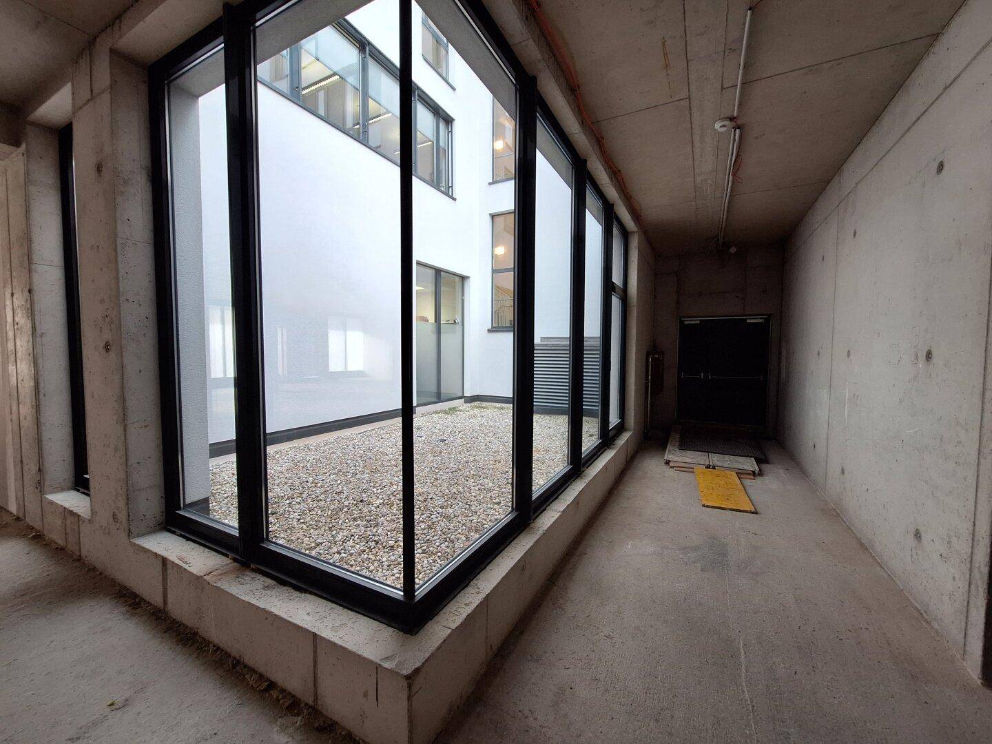 Innenraum mit großen Fenstern, die auf einen Kieshof blicken, umgeben von Betonwänden und -boden.