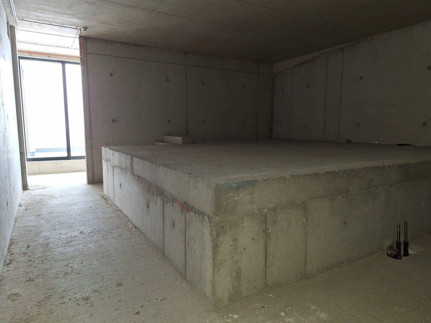 Unfertiger Raum mit einer erhöhten Betonplattform und einem Fenster im Hintergrund, ideal als Lagerbereich.