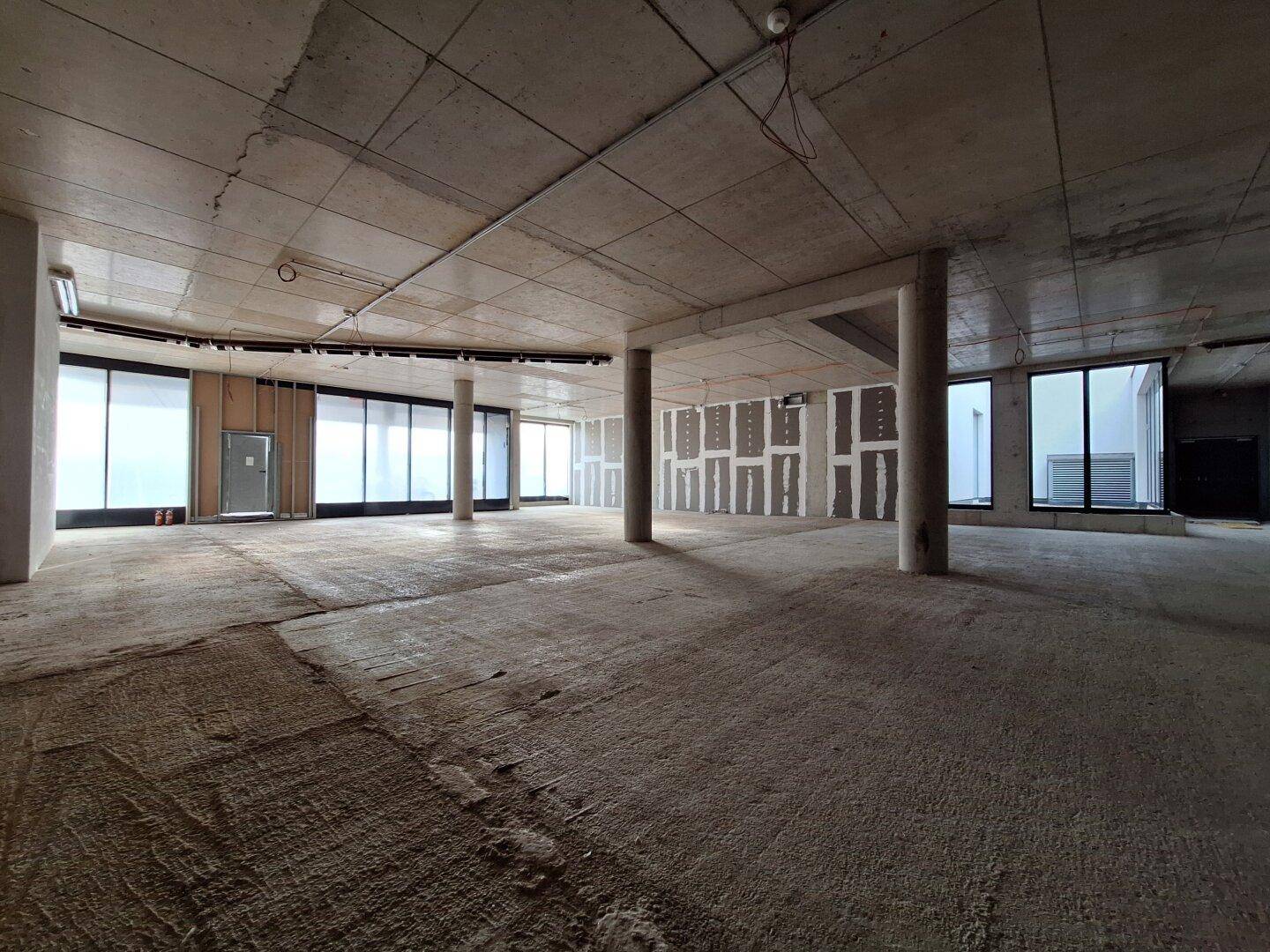 Weitläufiger Rohbau mit mehreren Betonsäulen und großen Fensterflächen, bietet viel Tageslicht und Raum.