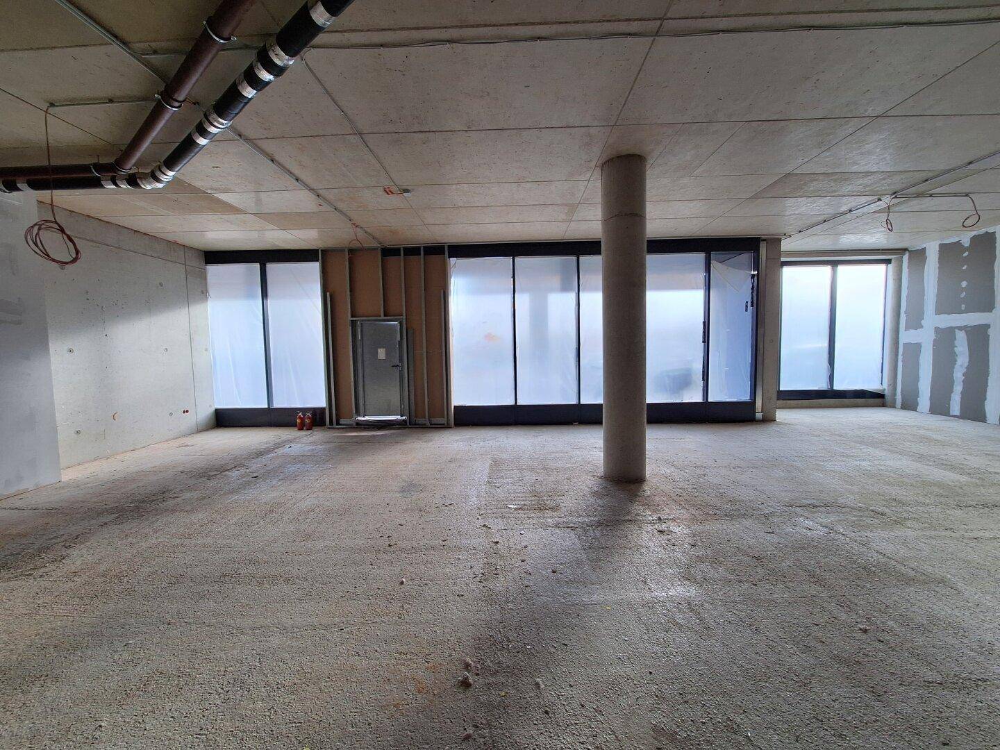 Rohbau mit großen, folierten Fensterfronten und einer Türöffnung, sichtbare Deckeninstallationen und Betonwände.