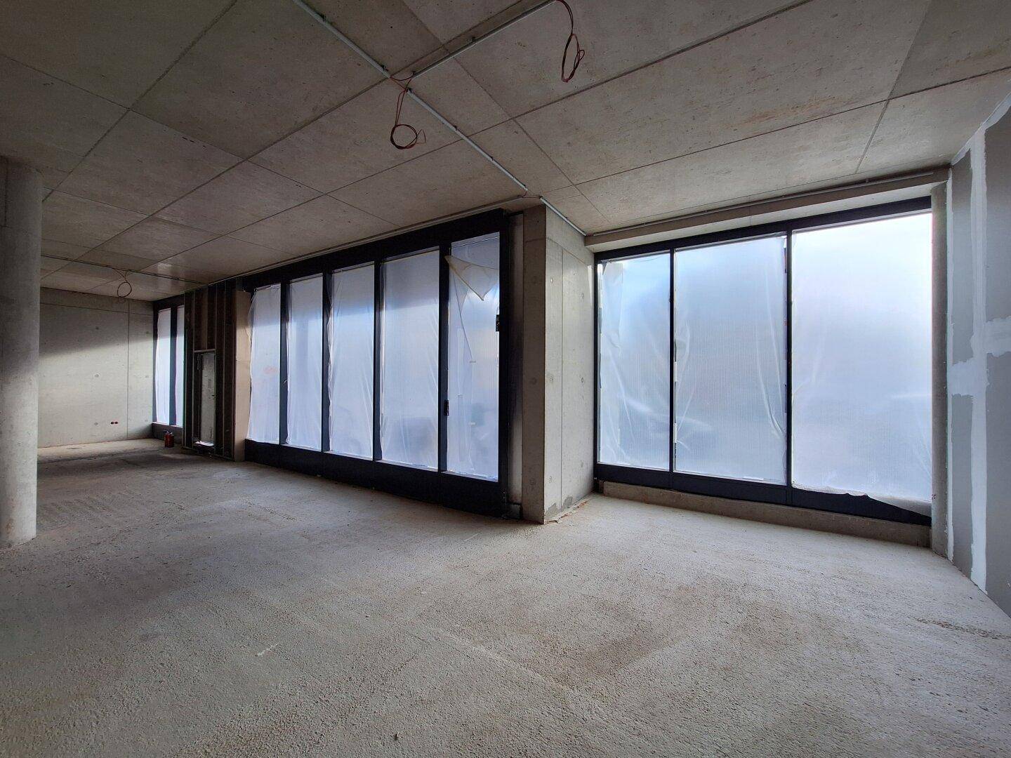 Geräumiger Rohbau mit großen, folierten Fensterfronten, Betonboden und -wänden, bietet viel Tageslicht.