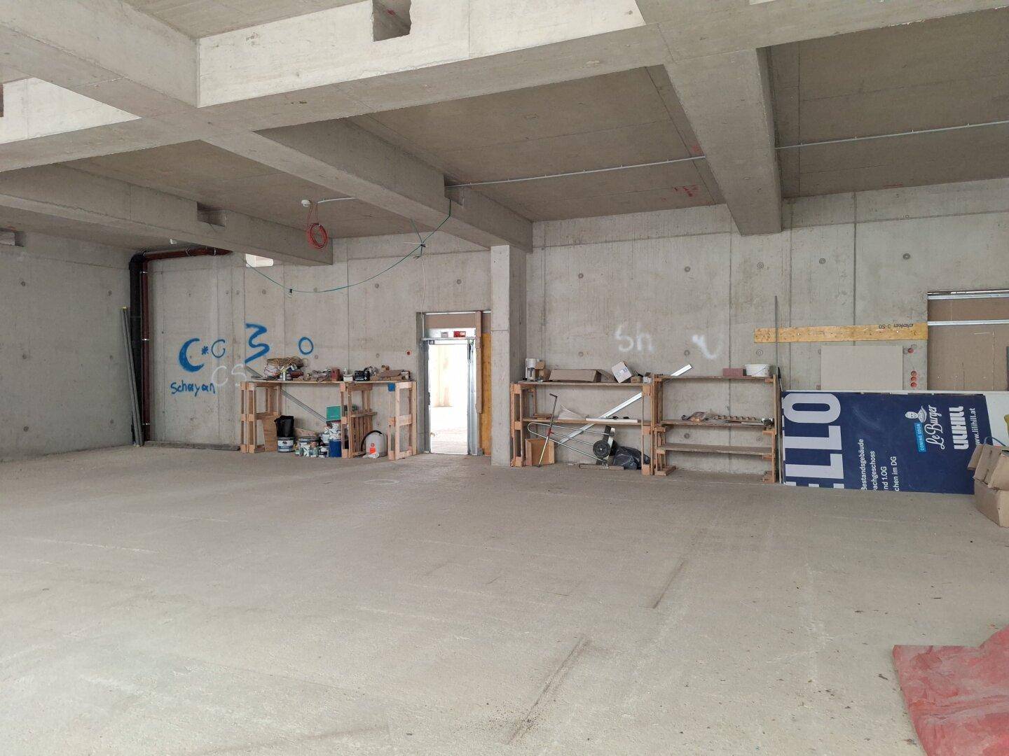 Rohbau mit sichtbaren Betonwänden, einigen Regalen und Spuren von Graffiti, ideal als Lager oder Werkstatt.