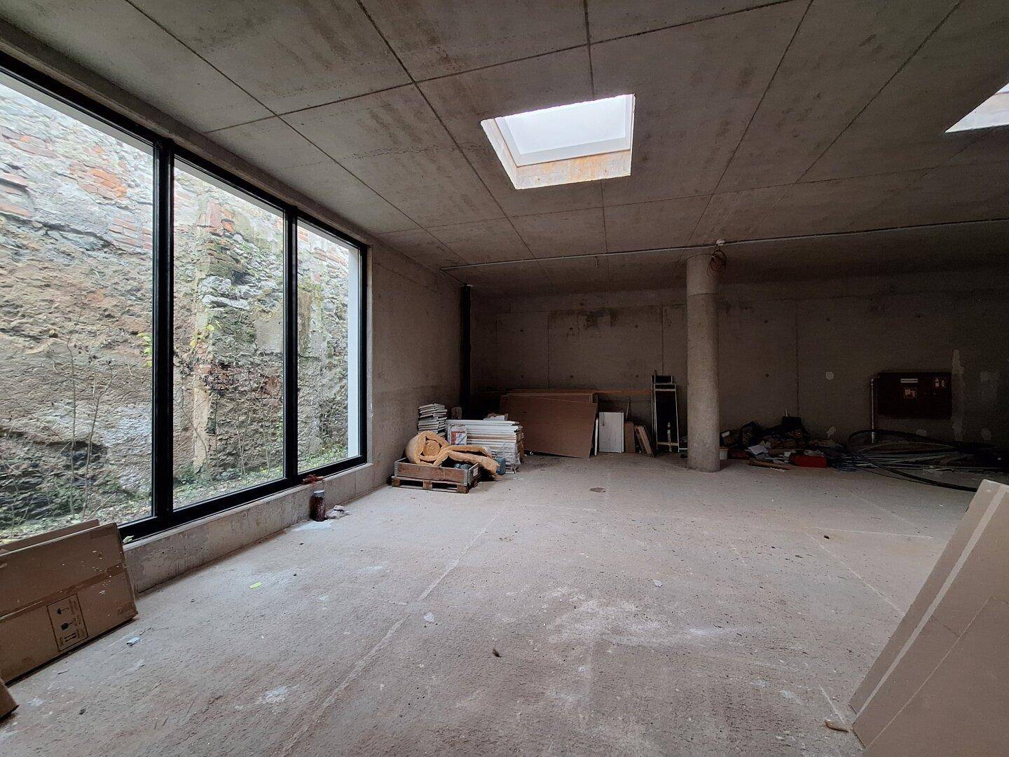 Rohbau mit großem Fenster zur Felswand und einem Oberlicht, Baumaterialien sind gestapelt.