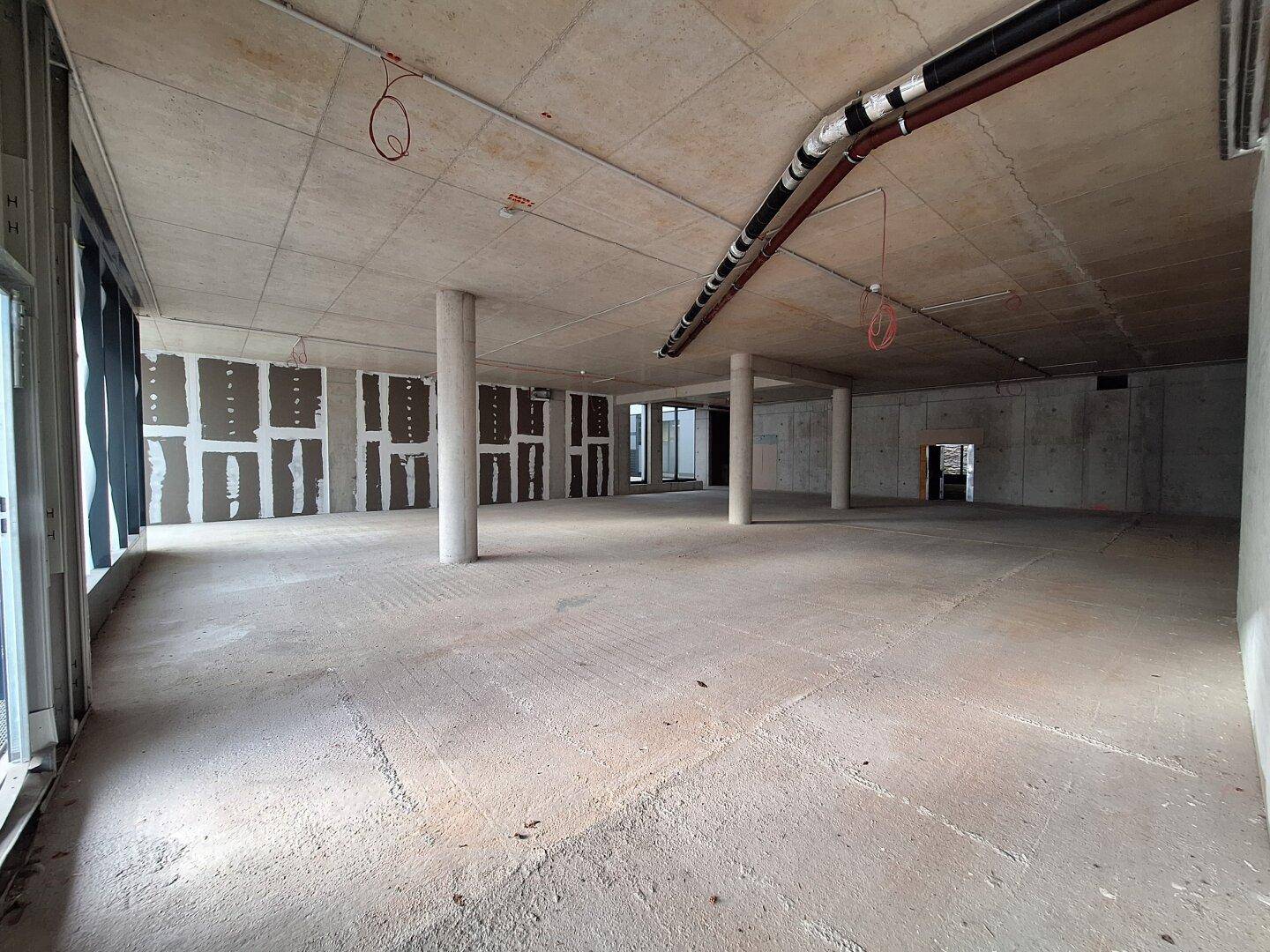 Geräumiger Rohbau mit sichtbaren Installationen, Betonsäulen und großen Fensterfronten, ideal für gewerbliche Nutzung.