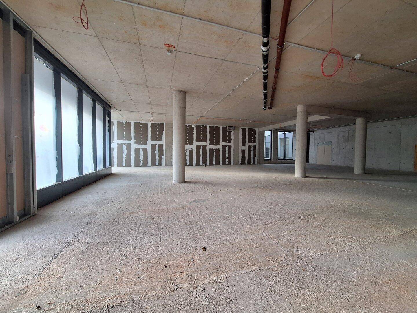Geräumiger Rohbau mit großen, folierten Fensterfronten, Betonsäulen und sichtbaren Deckeninstallationen.