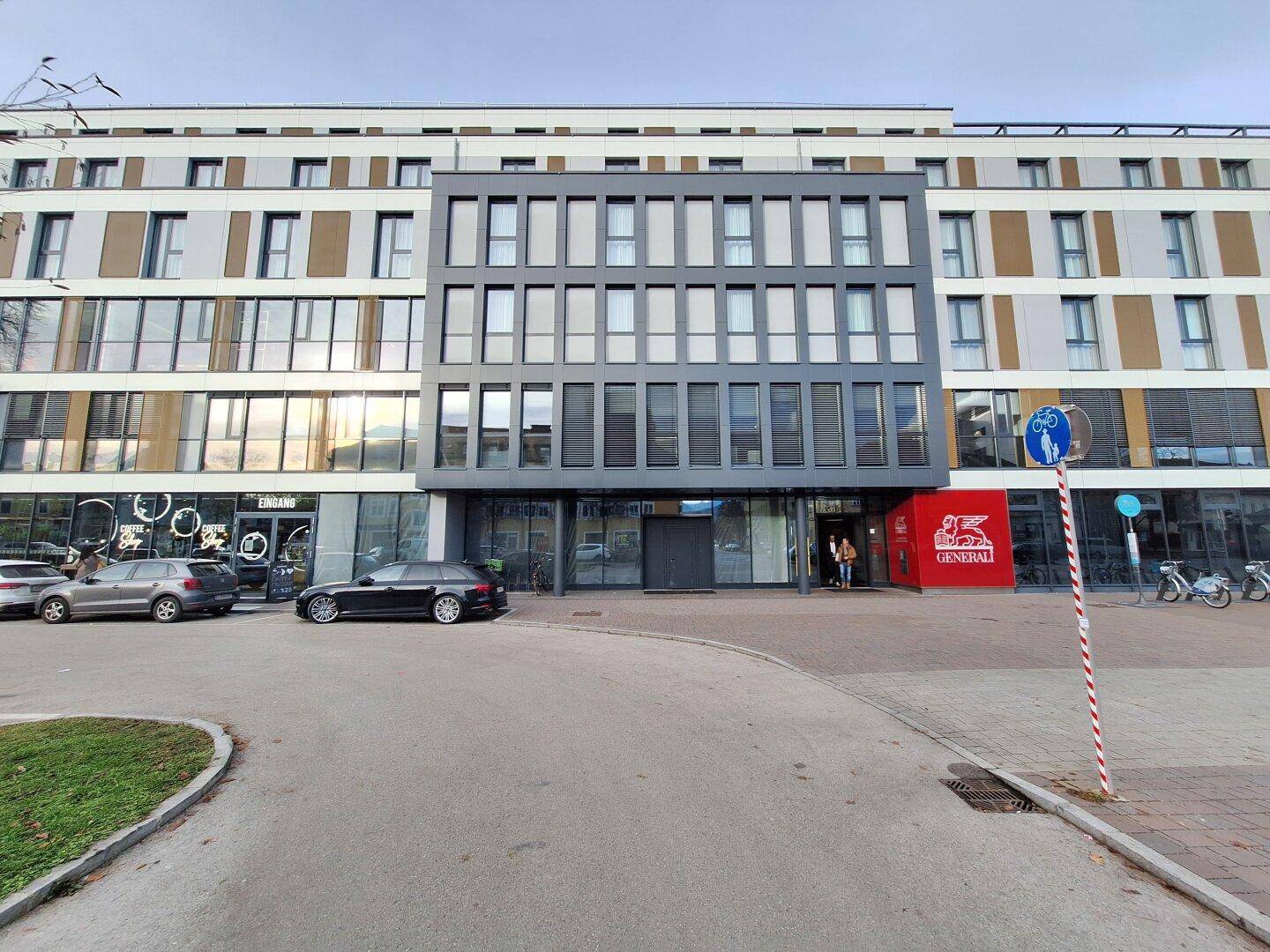 Moderne Gebäudefassade mit großen Fenstern und Geschäftsflächen im Erdgeschoss, umgeben von Parkplätzen.