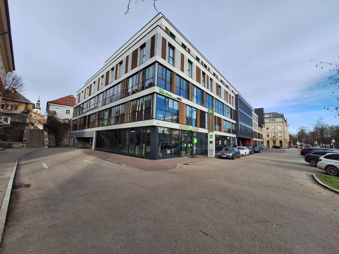 Weitwinkelansicht eines modernen Hotelgebäudes an einer Straßenecke mit umliegenden Gebäuden und Parkplätzen.