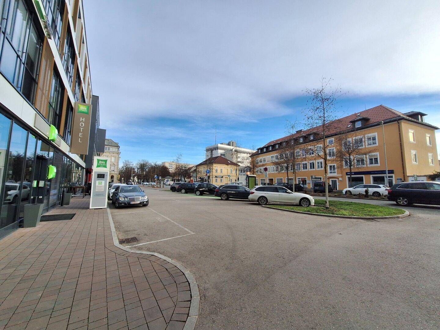 Straßenansicht mit modernem Hotelgebäude und geparkten Autos, umgeben von städtischer Architektur.