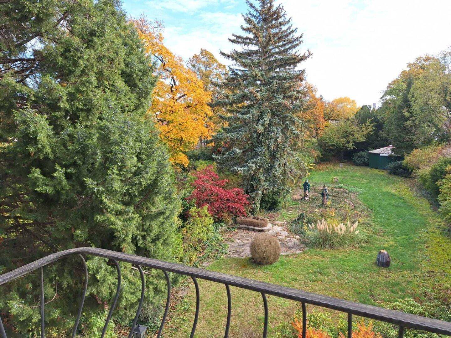 Weitläufiger Garten mit altem Baumbestand und herbstlicher Laubfärbung, von einem Balkon aus gesehen.