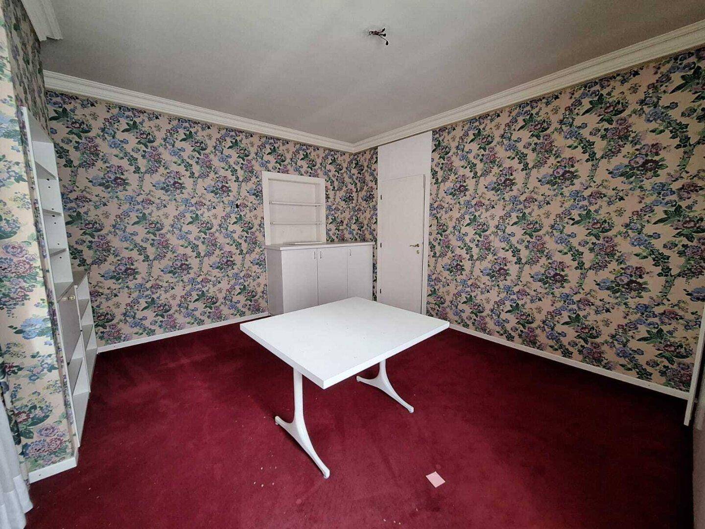 Schlafzimmer mit floraler Tapete, rotem Teppichboden und einem weißen Tisch, im Vintage-Stil.