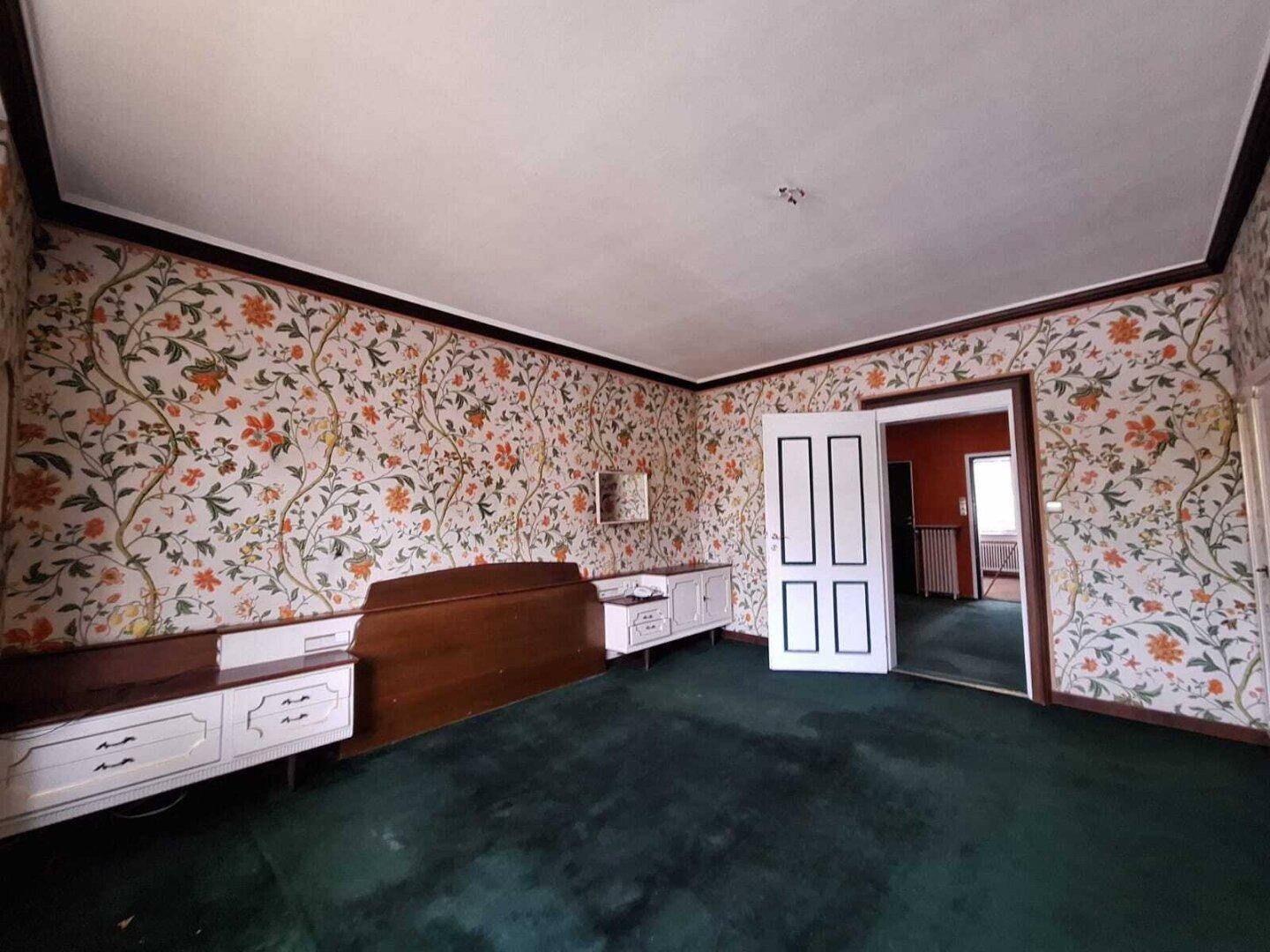 Schlafzimmer mit floraler Tapete, grünem Teppichboden und Einbauschränken, im Vintage-Stil.