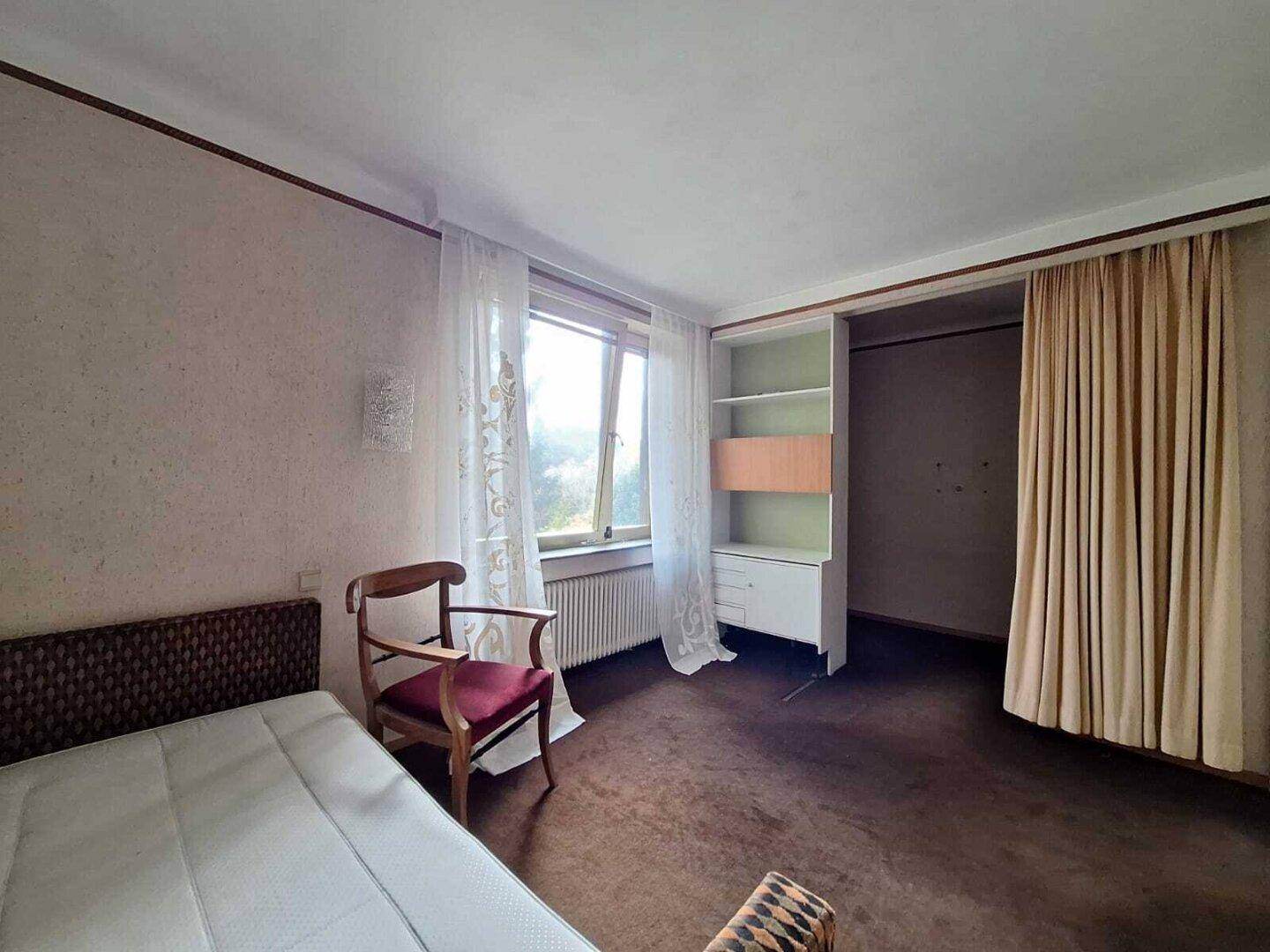 Schlafzimmer mit braunem Teppichboden, Fenster und einfacher Einrichtung, im Vintage-Stil.