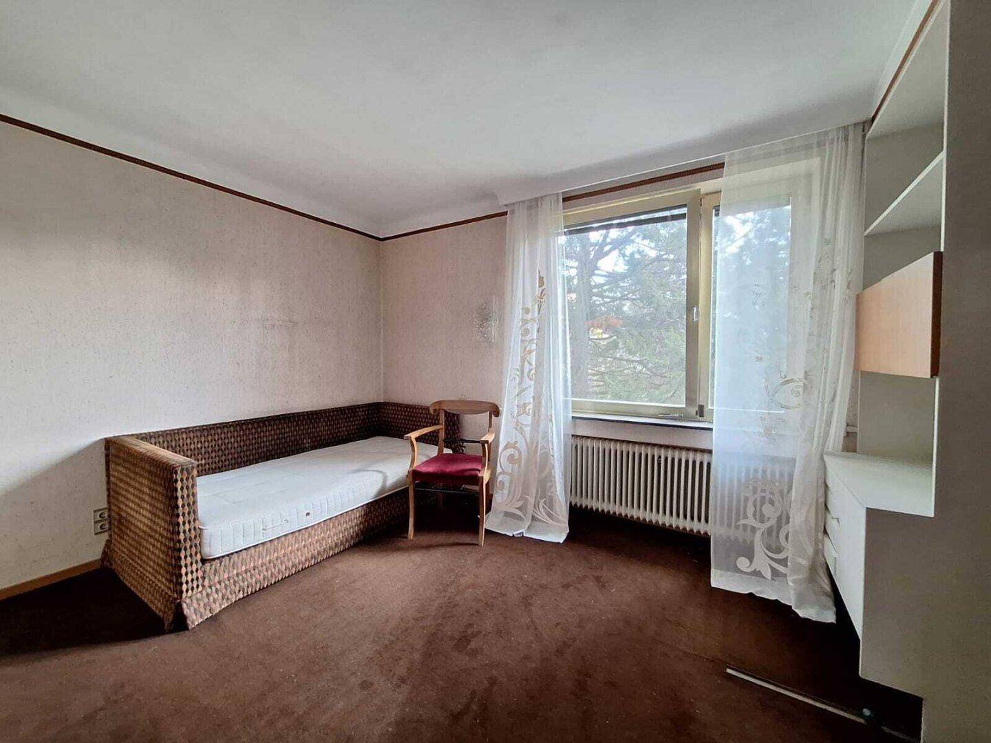 Schlafzimmer mit braunem Teppichboden, Fenster und einfacher Einrichtung, im Vintage-Stil.