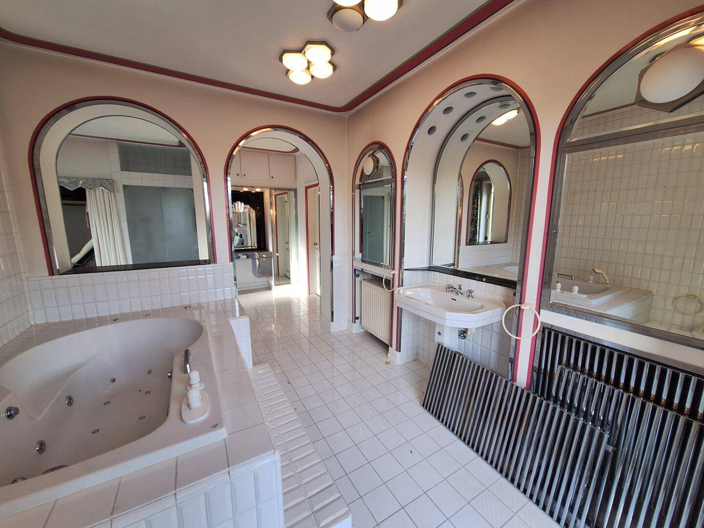 Großes Badezimmer mit Whirlpool-Badewanne, weißen Fliesen und mehreren Spiegeln im 80er-Jahre-Stil.