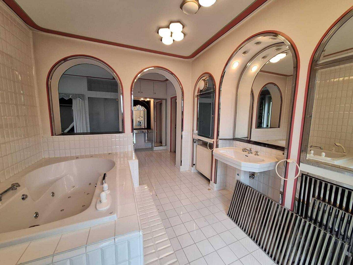 Großes Badezimmer mit Whirlpool-Badewanne, weißen Fliesen und mehreren Spiegeln im 80er-Jahre-Stil.