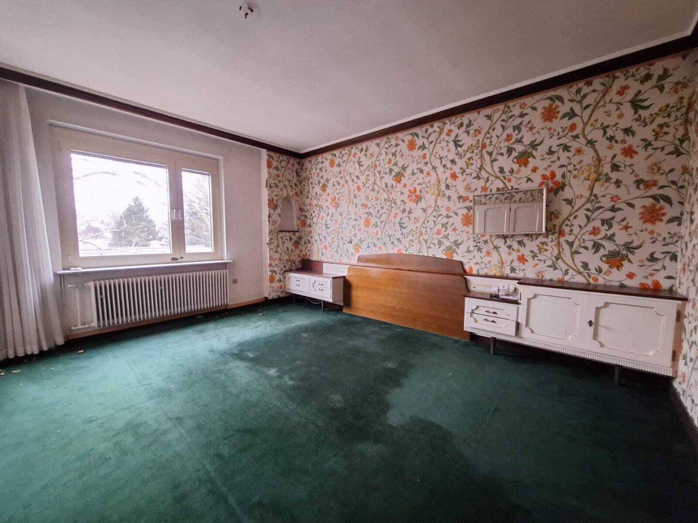 Schlafzimmer mit floraler Tapete, grünem Teppichboden und einem großen Fenster.