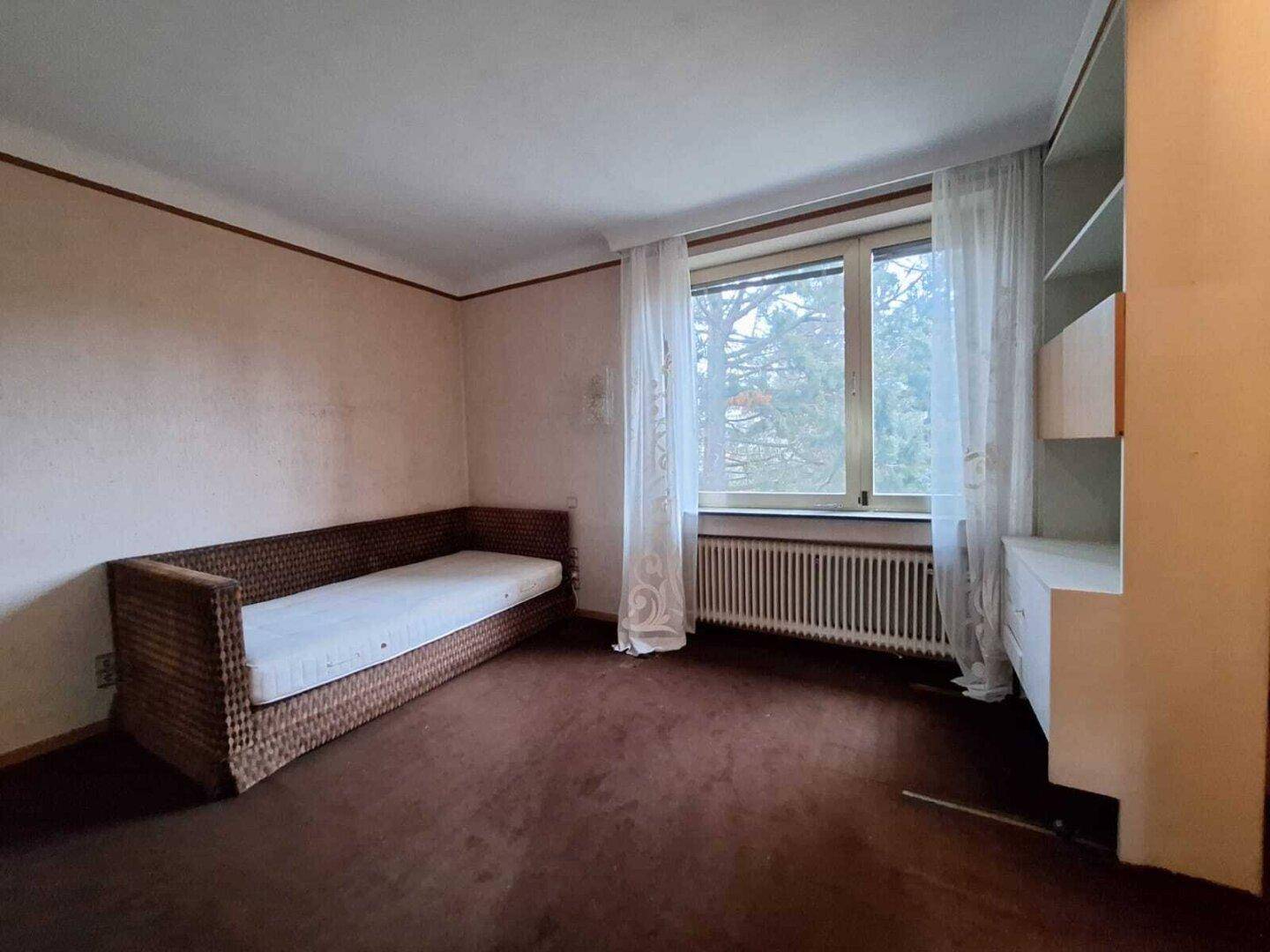 Helles Zimmer mit braunem Teppichboden, einem Tagesbett und einem großen Fenster.