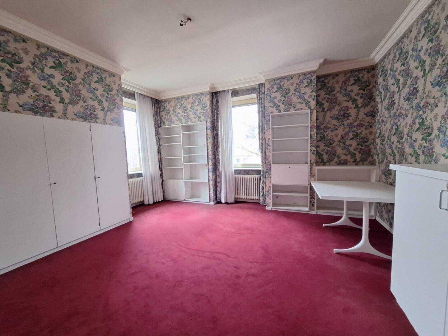 Schlafzimmer mit floraler Tapete, rotem Teppichboden und weißen Einbauschränken, im Vintage-Stil.
