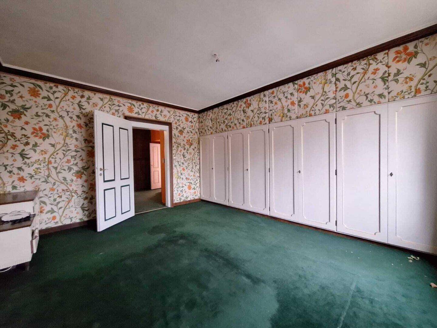 Schlafzimmer mit floraler Tapete, grünem Teppichboden und weißen Einbauschränken, im Vintage-Stil.