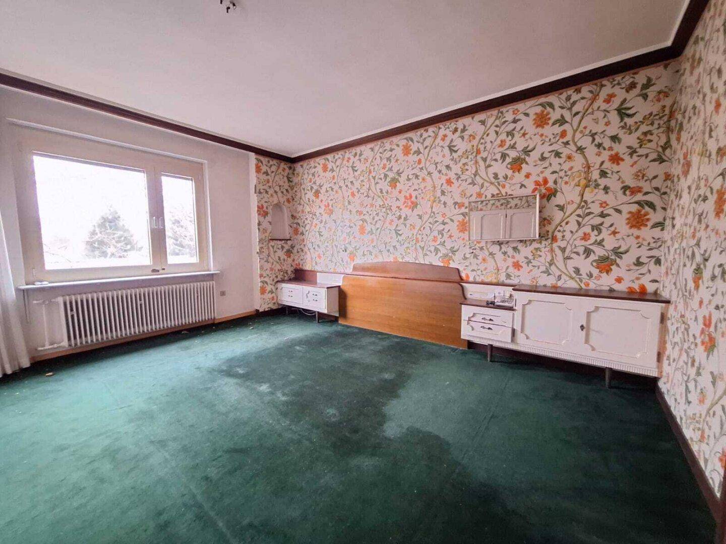 Schlafzimmer mit floraler Tapete, grünem Teppichboden und weißen Einbauschränken, im Vintage-Stil.