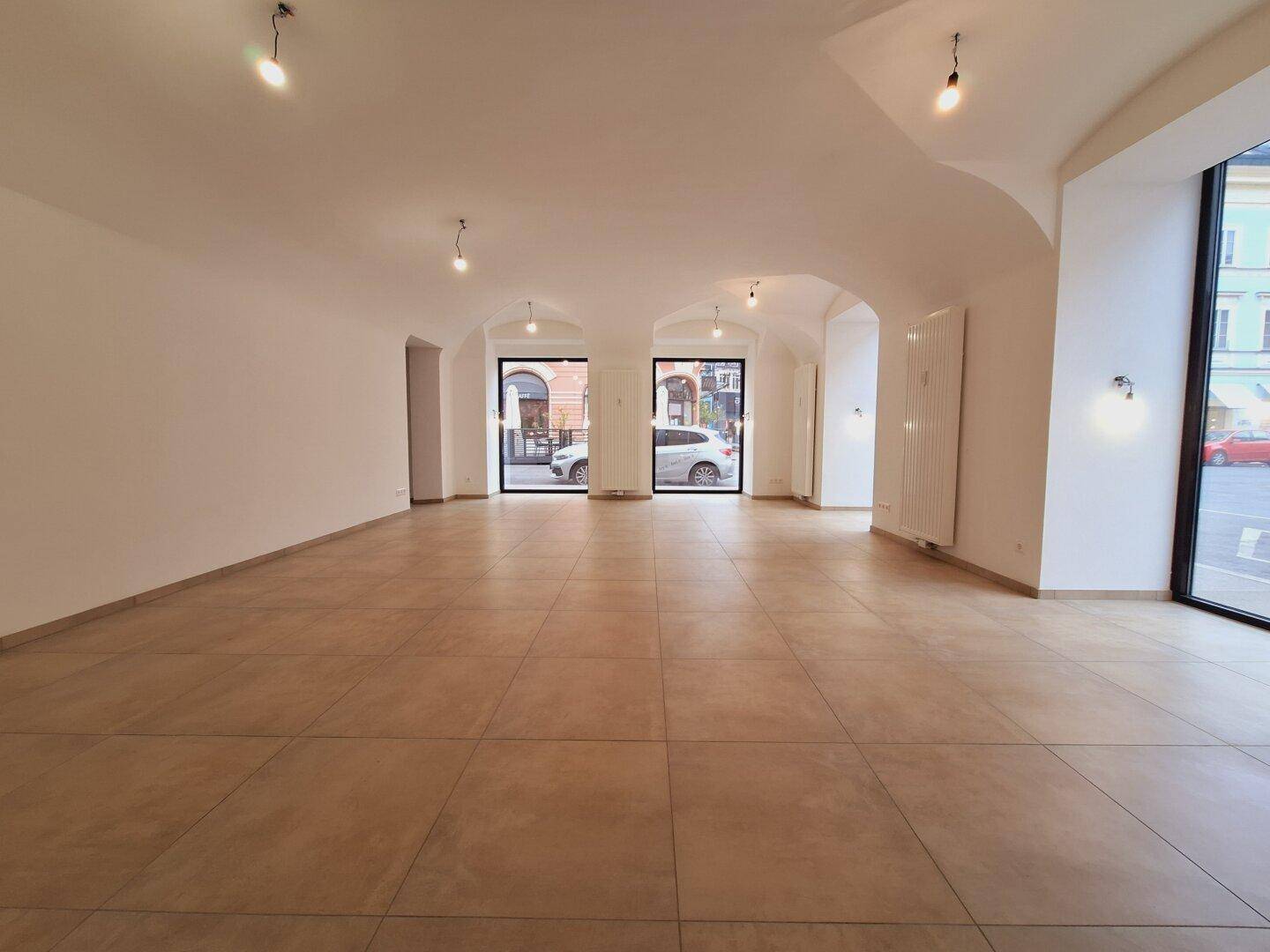 Heller Gewerberaum mit Fliesenboden und großen Fenstern, die viel Tageslicht hereinlassen und einen Blick auf die Straße bieten.