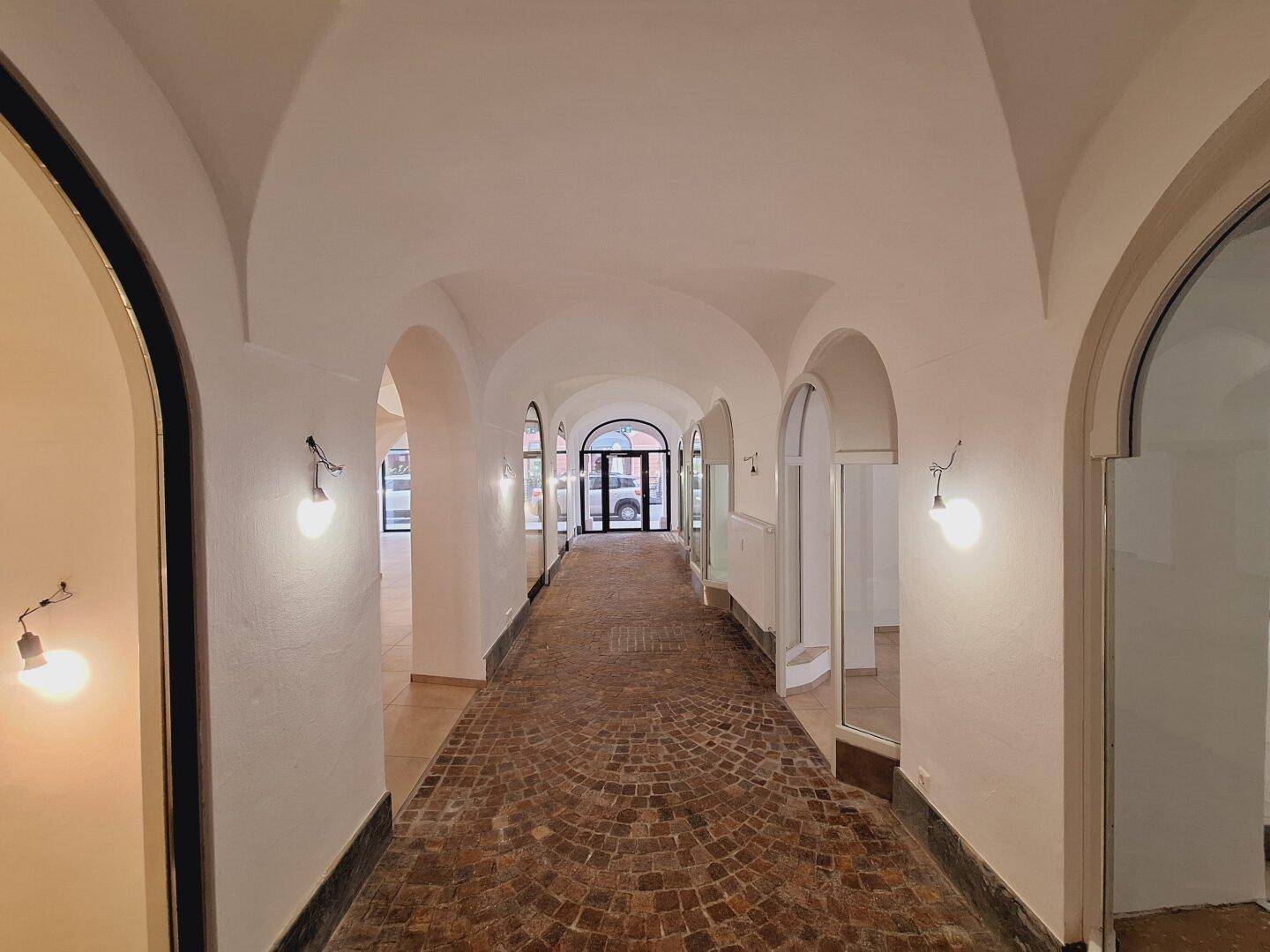Langer, gewölbter Durchgang mit Kopfsteinpflasterboden und beleuchteten Schaufenstern entlang der Seiten.