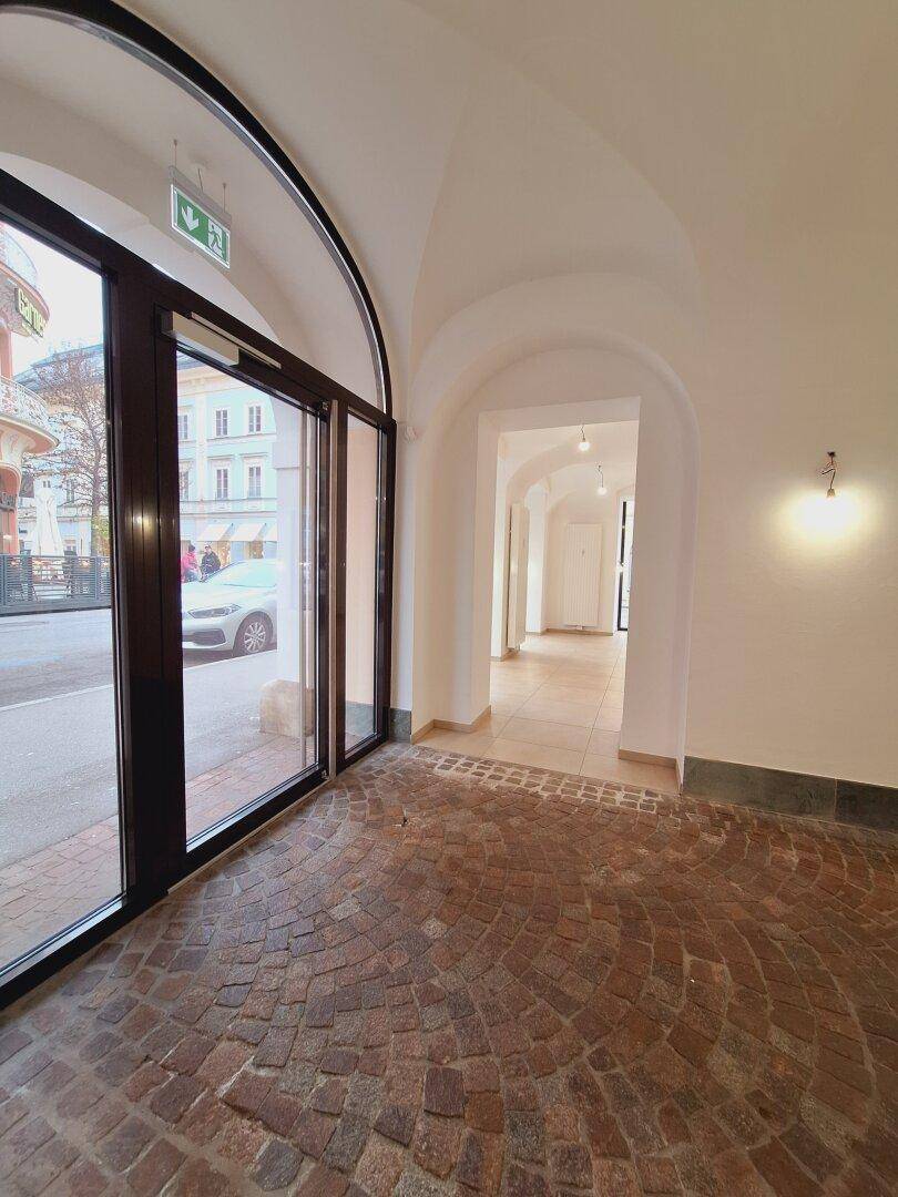 Eingangsbereich mit großen Glastüren, Kopfsteinpflasterboden und Blick in den gewölbten Durchgang.