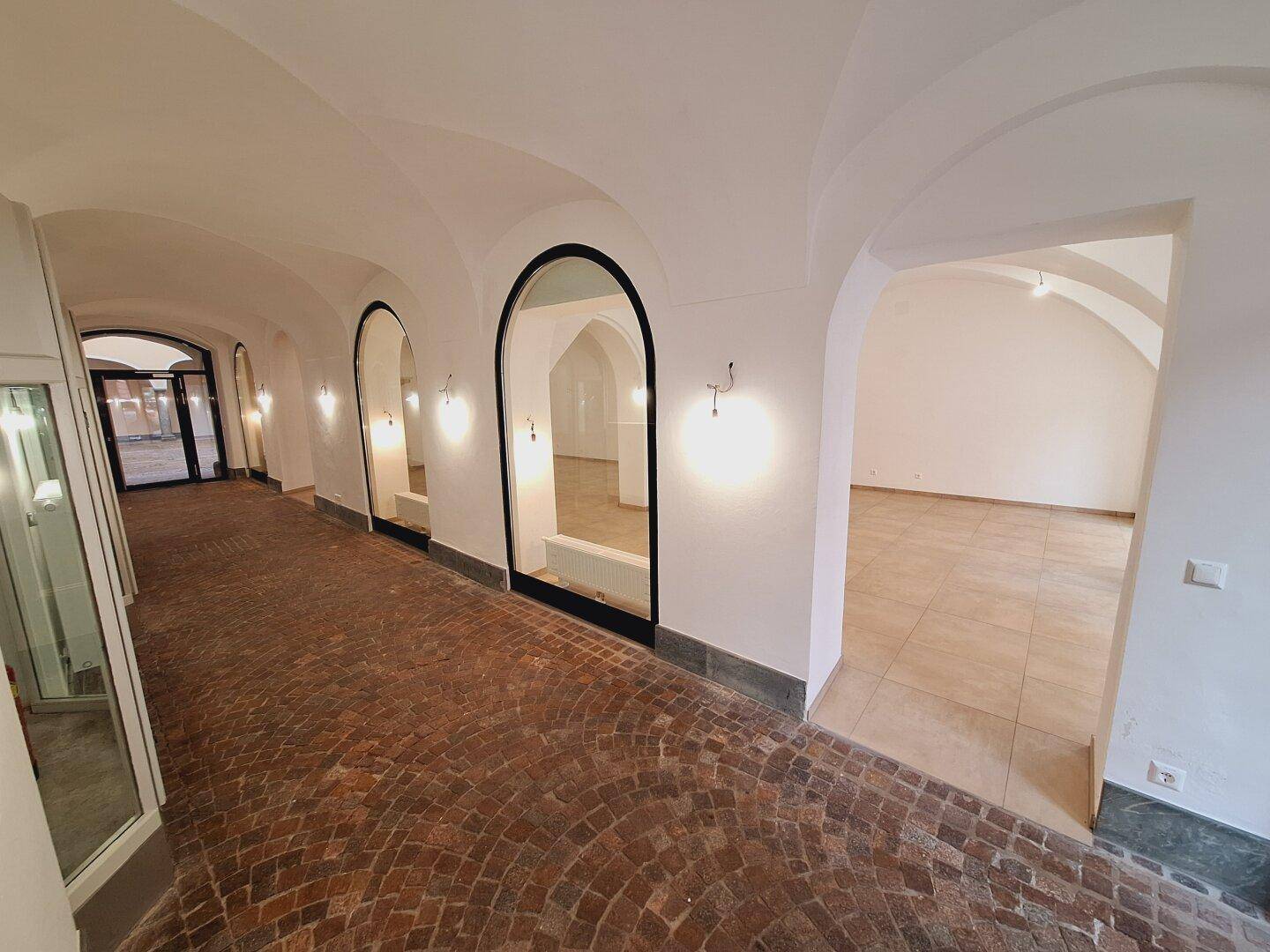 Gewölbter Durchgang mit Kopfsteinpflasterboden und Schaufenstern, der zu den Hauptgeschäftsflächen führt.